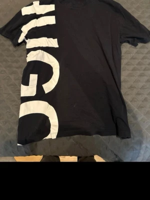 Svart HUGO t-shirt med stor logga - Snygg svart t-shirt från HUGO med stor vit logga längs vänster sida framtill. Klassisk passform och rund halsringning. Tillverkad i mjuk bomull som känns skön mot huden. Perfekt för dig som gillar streetwear och tydliga loggor.