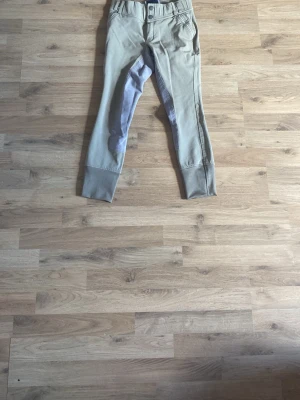 Beige ridbyxor med knäskoning - Snygga beige ridbyxor med förstärkt knäparti i grått och breda benslut. Byxorna har dubbla knappar i midjan och dragkedja. Perfekta för dig som gillar stilrena och funktionella ridkläder.