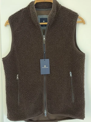 Brun pileväst från Hansen & Jacob - Mysig brun pileväst från Hansen & Jacob med hög krage och två dragkedjeförsedda fickor. Tillverkad i mjuk fleece med grov struktur och hel dragkedja framtill. Perfekt för lager-på-lager och en avslappnad stil.