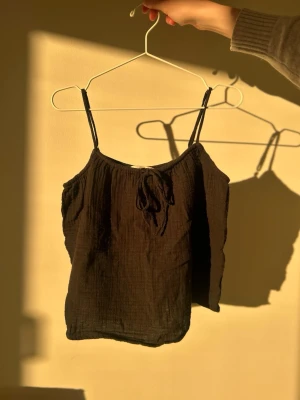 Svart linnetopp med knytband - En svart linnetopp med tunna axelband och knytband framtill. Toppen är tillverkad i ett lätt och luftigt bomullsmaterial med en avslappnad passform. Perfekt för varma dagar och enkel att matcha med jeans eller kjol.