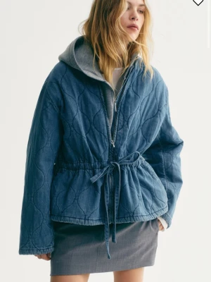 Oanvänd quiltad jeansjacka med lappen kvar - Säljer en blå quiltad jeansjacka med dragkedja och knytband i midjan. Jackan har rundad halsringning och långa ärmar. Perfekt för dig som gillar en avslappnad men ändå trendig look. Den är helt ny med prislappen kvar💕