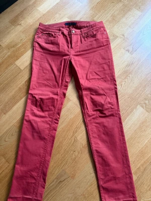 Röda jeans från Tommy Hilfiger - Snygga röda jeans från Tommy Hilfiger med klassisk femficksdesign och diskret logga på bakfickan. Jeansen har raka ben och är tillverkade i ett mjukt bomullsmaterial som ger en skön känsla. Perfekta för dig som vill sticka ut med färg.