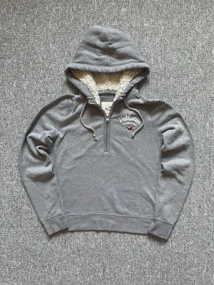 Grå hoodie från Hollister med foder - Mysig grå hoodie från Hollister med halv dragkedja och broderad logga på bröstet. Insidan av huvan är fodrad med fluffigt, vitt teddyfoder. Storleken är egentligen M men passar S.