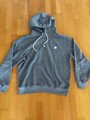 Acne studio hoodie - Snygg grå hoodie från Acne Studios med klassisk huva och vita dragsnören. Tröjan har en liten fyrkantig patch på bröstet och ribbade muddar vid ärmar och nederkant. Tillverkad i mjukt bomullsmaterial som känns skönt mot huden. Bara att höra av er vid minsta lilla fråga 😀