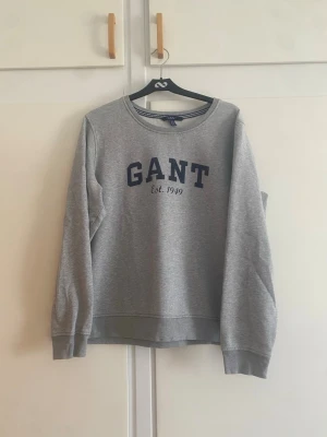 Grå sweatshirt från GANT - Klassisk grå sweatshirt från GANT med rund halsringning och blå logotyptext 'GANT Est. 1949' på bröstet. Tröjan har ribbade muddar vid ärmslut och nederkant. Perfekt för en avslappnad och stilren look.