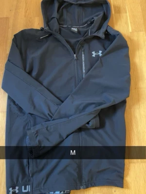 Svart vindjacka från Under Armour - Säljer en svart vindjacka från Under Armour med huva och dragkedja framtill. Jackan har logotyp på bröstet och diskreta detaljer längs dragkedjan. Perfekt för att hålla vinden borta under träning eller ute på stan.