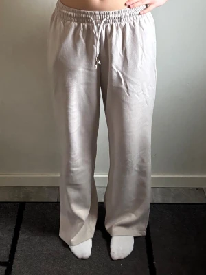 Beige mjukisbyxor med dragsko - Mysiga beige mjukisbyxor med rak passform och elastisk midja med dragsko. Perfekta för chill dagar och har en enkel, stilren look. Materialet är mjukt och skönt, vilket gör dem riktigt bekväma att ha på sig.