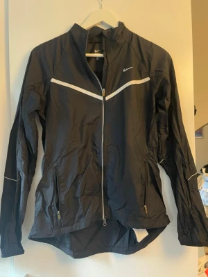 Svart vindjacka från Nike - Snygg svart vindjacka från Nike med vita detaljer och dragkedja framtill. Jackan har hög krage, långa ärmar och två fickor med dragkedja. Materialet är lätt och passar perfekt för utomhusaktiviteter. Nike-logga på bröstet och reflekterande detaljer.