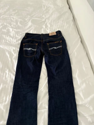 Mörkblå Nudie Jeans - Säljer ett par mörkblå jeans från Nudie Jeans med en slim/straight passform. Jeansen har kontrastsömmar i orange och vita broderade detaljer på bakfickorna. Tillverkade i kraftig denim. Jeansen är i storlek 27/32, men är lite uppsydda. Storleken motsvarar snarare 27/28.