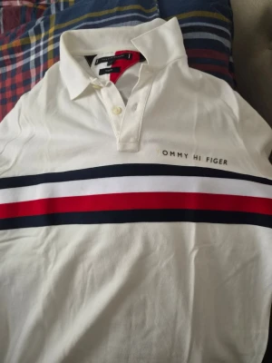 Vit slim fit skjorta från Tommy Hilfiger - Vit skjorta från Tommy Hilfiger i slim fit-modell med klassisk krage och knappar. Skjortan har breda horisontella ränder i blått, vitt och rött över bröstet samt logotyp på vänster sida. Materialet är bomull och designen är stilren och sportig.
