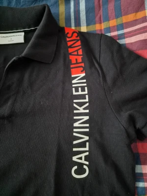 Svart pikétröja från Calvin Klein Jeans - Svart pikétröja från Calvin Klein Jeans med vit och röd logga längs ena sidan framtill. Slim fit-modell med klassisk krage och kort ärm. Perfekt för en stilren och modern look.