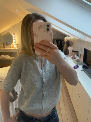 Ljusgrå kofta med kort ärm💗 - Storlek L men känns som xs/s🥰Mysig ljusgrå kofta med korta ärmar och knappar framtill. Tröjan har en mjuk, stickad känsla och en något croppad passform. Perfekt att slänga över ett linne eller t-shirt för en avslappnad look.