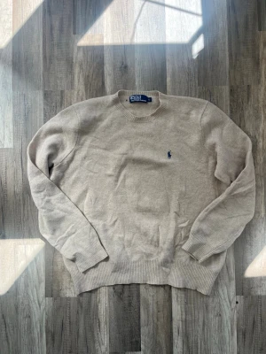 Beige stickad tröja från Polo Ralph Lauren - Säljer en beige stickad tröja från Polo Ralph Lauren med klassisk rund halsringning och blå broderad logotyp på bröstet. Tröjan har långa ärmar och ribbade muddar vid ärmslut och nederkant. Upplever att den är liten i storlek, så säljes på grund av för liten. Finns ett litet hål bredvid ettiketen. Tröjan är i lamm ull. 