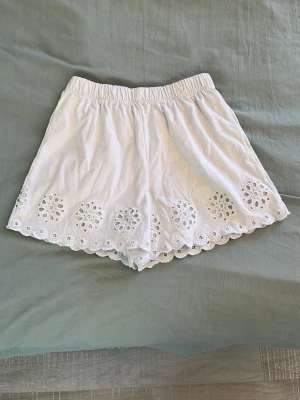 Vita broderade bomullsshorts - Säljer ett par vita shorts i bomull med elastisk midja och broderade detaljer längs nederkanten. Shortsen har en scallop-kant och ett luftigt, loose fit som passar perfekt till varma dagar. Broderierna ger en söt och unik touch.