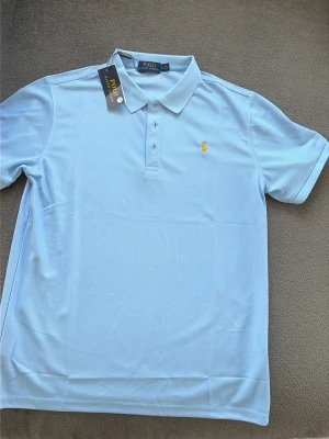 Ljusblå polotopp från Polo Ralph Lauren L - Snygg ljusblå polotopp från Polo Ralph Lauren med klassisk krage och tre knappar framtill. T-shirten har korta ärmar och ett broderat gult logomärke på bröstet. Perfekt för dig som gillar stilrena och tidlösa plagg.