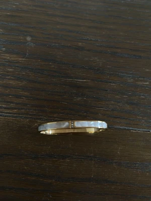 Guldigt armband med pärldetaljer - Snyggt armband i guldfärgad metall med vita pärldetaljer längs hela bandet. Armbandet har en rund form och stilren design som passar perfekt för dig som gillar klassiska accessoarer med en twist.