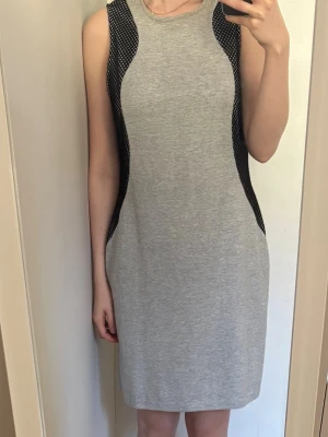 Grå ärmlös klänning med nätmönster - Snygg grå ärmlös klänning med svarta nätmönstrade paneler på sidorna som ger en sportig och modern look. Klänningen har dragkedja baktill .