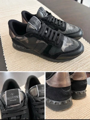 Valentino Garavani svarta sneakers - Snygga svarta sneakers från Valentino Garavani med kamouflagemönster och coola nitar på sulan. Skorna har en mix av mocka och läder, samt är dem använda med milda deffekter, som att det är släppt lite på vissa ställen, metalliska detaljer vid hälen. Perfekta för dig som vill ha en unik och trendig look.