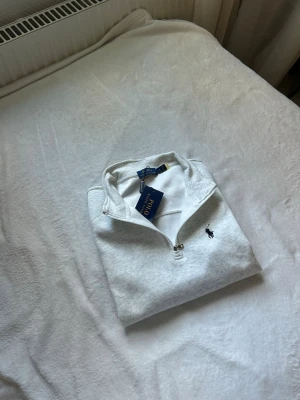 Vit half-zip tröja från Polo Ralph Lauren - Snygg vit half-zip tröja från Polo Ralph Lauren med klassisk blå logga broderad på bröstet. Tröjan har hög krage och dragkedja framtill, perfekt för en clean och stilren look. Tillverkad i mjukt material för extra komfort.