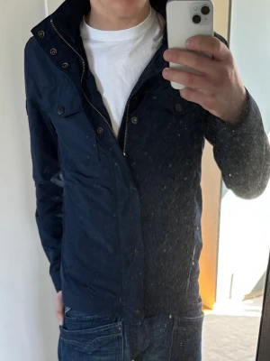 Marinblå fieldjacka från J.Lindeberg - Snygg marinblå fieldjacka från J.Lindeberg med dragkedja och tryckknappar framtill. Jackan har hög krage, två bröstfickor med lock och ett diskret JL-märke på ärmen. Perfekt för dig som vill ha en stilren och funktionell jacka.