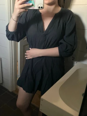 Svart kort klänning med v-ringning - En svart kort klänning med v-ringad hals och trekvartsärmar. Klänningen har en lös passform och är tillverkad i ett lätt, skrynkligt tyg som ger en avslappnad känsla. Perfekt för dig som gillar enkel och stilren design.