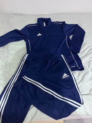 Marinblå Adidas träningsset - Säljer ett marinblått träningsset från Adidas med långärmad tröja och matchande byxor. Setet har vita detaljer och klassiska Adidas-ränder längs benen samt logga på både tröja och byxor. Materialet är mjukt och stretchigt, perfekt för träning eller chill.