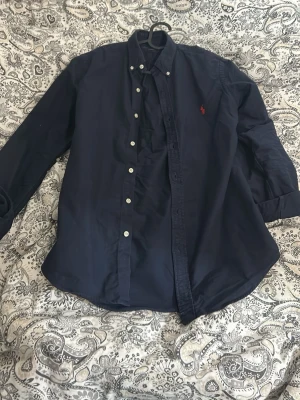 Mörkblå skjorta från Ralph Lauren - Säljer en mörkblå skjorta från Ralph Lauren med slim fit-passning. Skjortan har klassisk krage, vita knappar och det ikoniska röda Polo-logotypen broderad på bröstet. Perfekt för dig som gillar stilrena och tidlösa plagg. Pris går att diskutera, har inga defekter och är i väldigt bra skick!