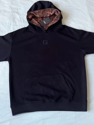 Svart hoodie från Fendi med FF-mönster - Svart hoodie från Fendi med klassisk känguruficka och broderad Fendi-logga på bröstet. Insidan av huvan har det ikoniska bruna FF-mönstret. Tillverkad i mjukt material som känns lyxigt och bekvämt. Perfekt för dig som gillar exklusiv streetwear.