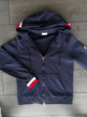 Mörkblå hoodie från Moncler - Snygg mörkblå hoodie från Moncler med dragkedja, huva och fickor framtill. Ärmsluten har coola detaljer i rött, vitt och grått. Moncler-logga på ena ärmen. Materialet är mjukt och bekvämt, perfekt för chill dagar.