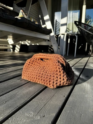 Brun virkad clutch bag  - Clutch väska i brun färg, virkad med tjockt garn för en chunky och mjuk känsla. Väskan har en rektangulär form och ett integrerat handtag upptill. Insytt teal blått silkes foder. Materialet är bomull och designen är egengjord, vilket ger en unik och personlig touch till din stil. Priset är satt efter material och tid.