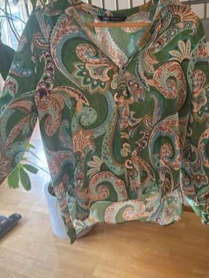 Grön paisleymönstrad blus från Zara - Snygg grön blus från Zara med paisleymönster i turkos, beige och brunt. Blusen har v-ringning och långa ärmar. Materialet är lätt och luftigt, perfekt för att skapa en cool och avslappnad look.