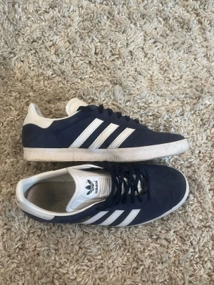 Adidas Gazelle sneakers marinblå - Snygga Adidas Gazelle sneakers i marinblå mocka med klassiska vita ränder och vit häl. Skorna har snörning och vit sula, perfekt för dig som vill ha en stilren och ikonisk look. Adidas-logga på tungan och hälen. skicket är 8/10 lite täcken på andningen men går lätt o tvätta bort. ingen box tillkommer 
