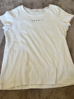 Vit t-shirt från Esprit med nitar - Vit t-shirt från Esprit med rund halsringning och korta ärmar. Framtill finns Esprit-loggan dekorerad med små silverfärgade nitar för en stilren och enkel look. Tillverkad i mjuk bomull som känns skön mot huden.