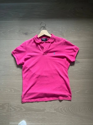 Rosa pikétröja från Polo Ralph Lauren - Klassisk rosa pikétröja från Polo Ralph Lauren med liten blå logga broderad på bröstet. Tröjan har korta ärmar, krage och knappar vid halsen. Tillverkad i 100% bomull för en skön och luftig känsla.