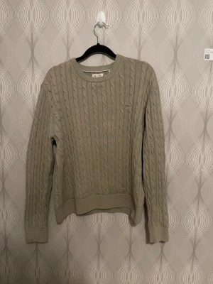 Lite grön Beige kabelstickad tröja från Gant - Säljer en lite grön beige kabelstickad tröja från Gant med rund hals och diskret logotyp på bröstet. Tröjan har ribbade muddar vid ärmslut och nederkant, och är gjord i mjukt material som passar perfekt för lager-på-lager. Klassisk och stilren design.
