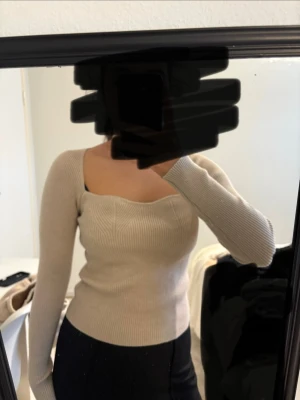 Beige stickad topp med sweetheart-urringning - Säljer en beige stickad topp med långärmad design och snygg sweetheart-urringning. Tröjan är figurnära och har ribbstickat material som ger en cool och modern look. Perfekt för dig som vill ha något stilrent och trendigt.