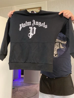 Svart sweatshirt från Palm Angels - Säljer en svart sweatshirt från Palm Angels med stor vit logga och text på bröstet. Tröjan har rund halsringning, långa ärmar och breda muddar vid ärmslut och nederkant. Perfekt för dig som gillar streetwear och vill sticka ut med en ikonisk design.