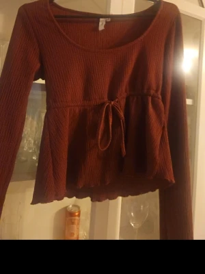 Vinröd ribbad topp med knyt - Säljer en vinröd långärmad topp med ribbad struktur och knyt i midjan. Toppen har en rund halsringning och volangdetalj nedtill, vilket ger en snygg och avslappnad look. Perfekt för dig som gillar trendiga och bekväma plagg.