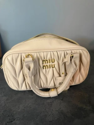 Beige Miu Miu väska - Säljer en beige handväska från Miu Miu, köpte här på plick så osäker om den är 100% äkta. Men känns väldigt äkta, har bra kvalité och der nästan helt ny ut. Säljer för att jag samlar ihop till en ny väska. Lägg gärna bud