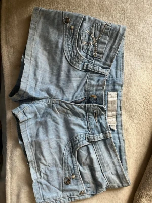 Ljusblå denimshorts med paljettdetaljer - Säljer ett par ljusblå denimshorts med snygga paljettdetaljer och kedjor på bakfickorna. Midjemått ungefär 35 cm. Perfekta för sommaren och ger en cool touch till din outfit. Hör av hid frågor och pris kan diskuteras!💗