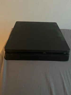 PlayStation 4 Slim svart konsol - Säljer en svart PlayStation 4 Slim från Sony. Konsolen har en stilren och platt design med mattsvart finish och PlayStation-loggan på ovansidan. Perfekt för gaming och streaming, tillverkad i plast.
