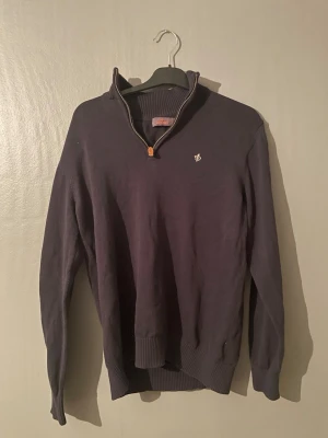 Mörkblå halvzip-tröja från Morris - Mörkblå stickad tröja från Morris med halvzip och hög krage. Tröjan har ett diskret broderat logomärke på bröstet och ribbade muddar vid ärmslut och nederkant. Perfekt för lager-på-lager och snygg till jeans.