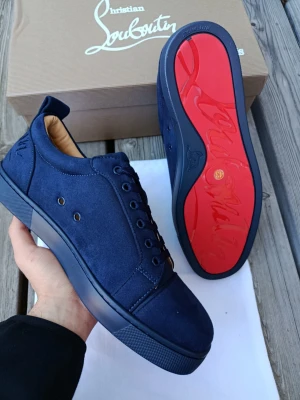 Blåa sneakers från Christian Louboutin - Snygga blåa sneakers från Christian Louboutin med klassisk röd sula och ovandel i mocka. Skorna har diskreta detaljer, ton-i-ton snörning och metallöglor på sidan. Perfekt för dig som vill ha en exklusiv och stilren look.