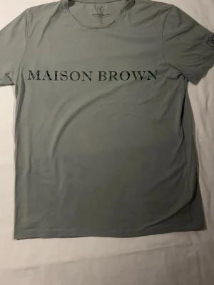 Grå t-shirt från Maison Brown - Snygg grå t-shirt från Maison Brown med klassisk rund hals och tryck i svart text på bröstet. T-shirten har korta ärmar och är tillverkad i mjukt bomullsmaterial som känns skönt mot huden. Perfekt för dig som gillar stilrena plagg med diskret branding.