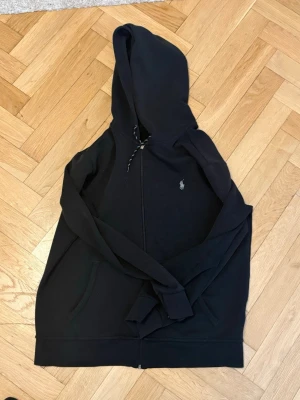 Svart hoodie från Polo Ralph Lauren - Säljer en svart hoodie från Polo Ralph Lauren med dragkedja framtill och klassisk huva. Tröjan har fickor på sidorna och den ikoniska lilla logotypen broderad på bröstet. Perfekt för en avslappnad och stilren look.