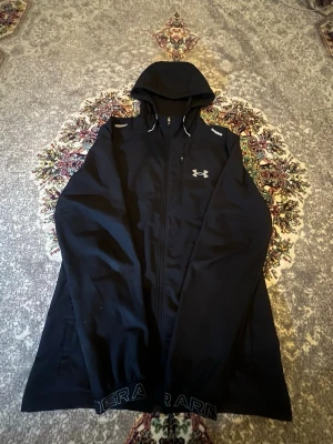 Under armour jacka - Snygg svart vindjacka från Under Armour med huva och dragkedja framtill. Jackan har logotyp på bröstet, reflexdetaljer på axlarna och fickor med dragkedja. Materialet är lätt och sportigt, perfekt för aktiva dagar.