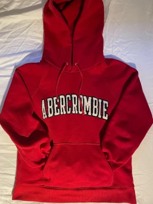 Abercrombie & fitch Vintage - Det står M men passar Xs-S. Säljer nu denna supersnygga och klassiska hoodie från Abercrombie & Fitch i en riktigt härlig röd färg! Tröjan har den ikoniska broderade loggan över bröstet i vitt och marinblått, samt en liten logga på huvan. Den har en perfekt vintage/Y2K-vibe och är gjord i ett rejält, mjukt material som håller värmen bra. Raglanärmarna ger den en snygg passform. • Skick: Mycket fint begagnat skick, väl omhändertagen! • Färg: Klarröd med vita detaljer.