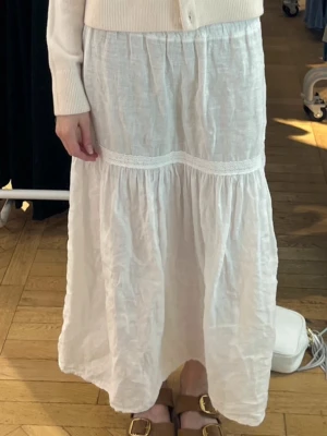 Vit långkjol i linne med spetsdetalj - Säljer en vit långkjol i luftigt linne med en fin spetsdetalj vid höften. Kjolen har en avslappnad passform och faller snyggt mot benen. Perfekt för dig som gillar en enkel men stilren look. Ingen storleklapp men skulle säga att den passar till en xs-s