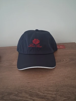 Mörkblå keps från Loro Piana - Snygg mörkblå keps från Loro Piana med röd broderad logga och text framtill. Kepsen har böjd skärm med vit kant och justerbar resår baktill. Tillverkad i ett slätt, lätt material som ger en clean och sportig look.