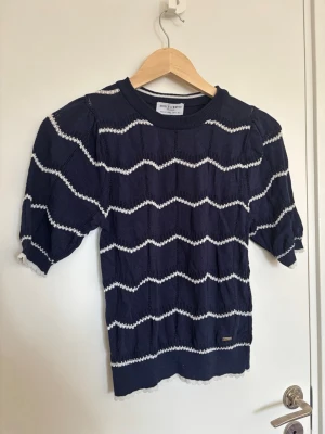Mörkblå stickad topp från Holly & Whyte by Lindex - Snygg mörkblå stickad topp från Holly & Whyte by Lindex med vita vågiga ränder. Modellen har korta puffärmar och rund halsringning. Materialet är mjukt och lätt, perfekt för lager på lager. Fina detaljer med vit kant vid ärmslut och nederkant.